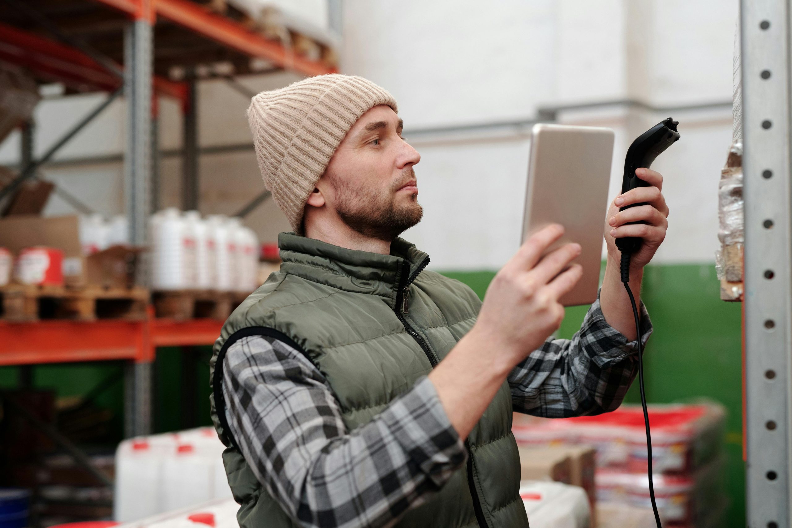 Man in beanie and vest using a scanner in a warehouse for inventory control.